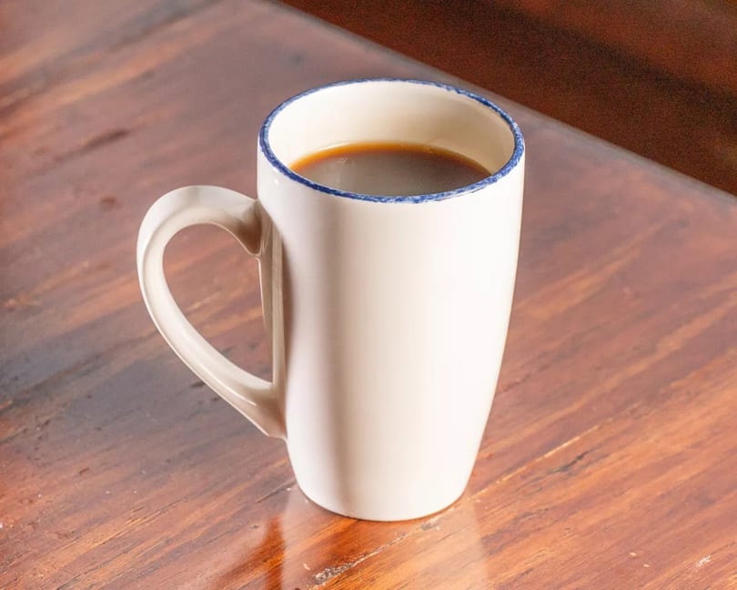 White ceramic mug with blue rim, filled with dark coffee, on a wooden table.