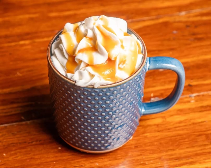 A blue textured mug filled with whipped cream and drizzled with caramel sauce on a wooden table.