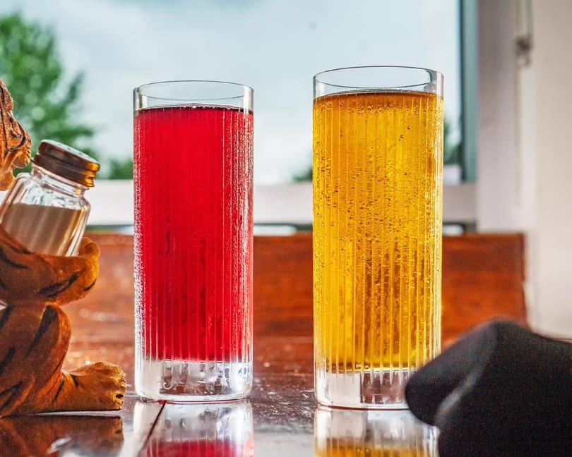 Two tall glasses, one with red and one with yellow bubbly drinks, on a wooden table.