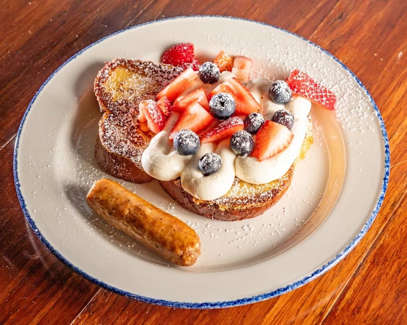 French toast topped with whipped cream, strawberries, blueberries, powdered sugar, and a sausage link.