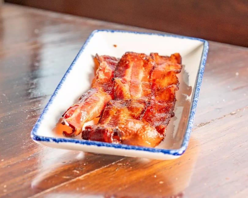 Crispy, glazed bacon strips served in a white rectangular dish with blue trim on a wooden table.