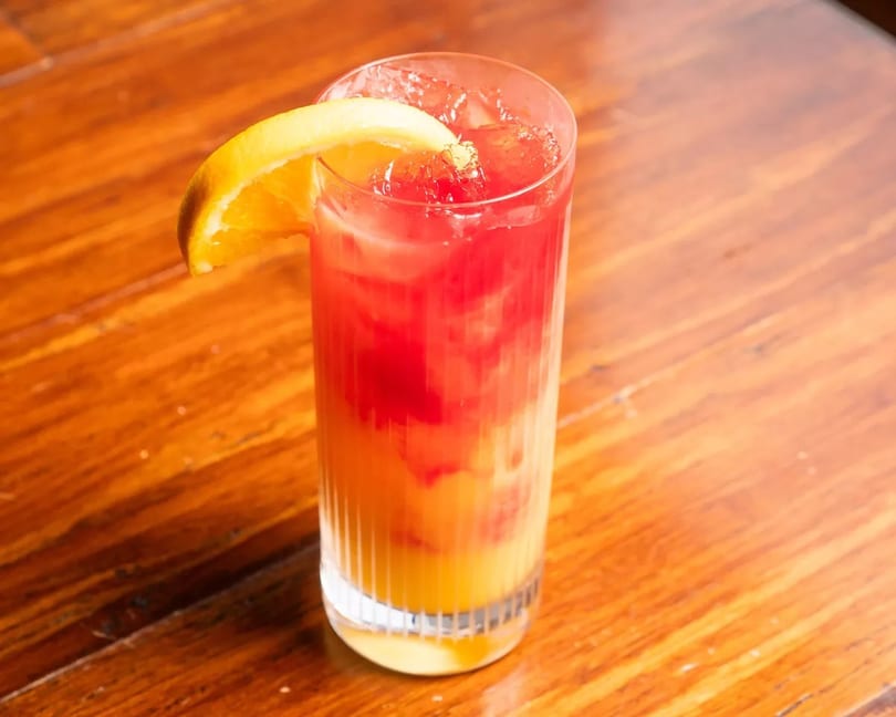 Tall glass with a layered red and orange drink, garnished with an orange slice on a wooden table.